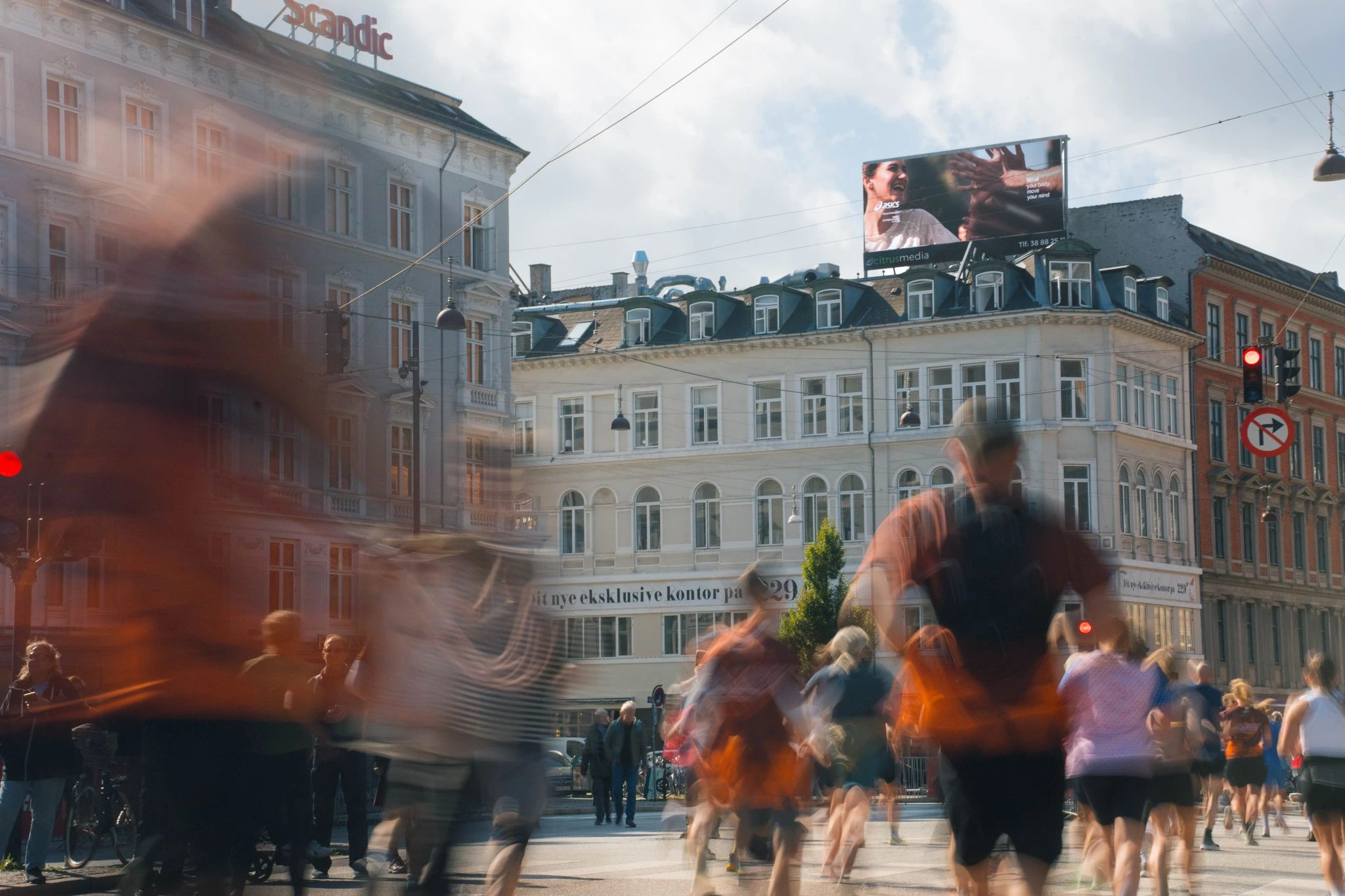 Billboard de ASICS visible sobre un edificio de Copenhague mientras corredores y peatones se desplazan por la calle durante una activación urbana.