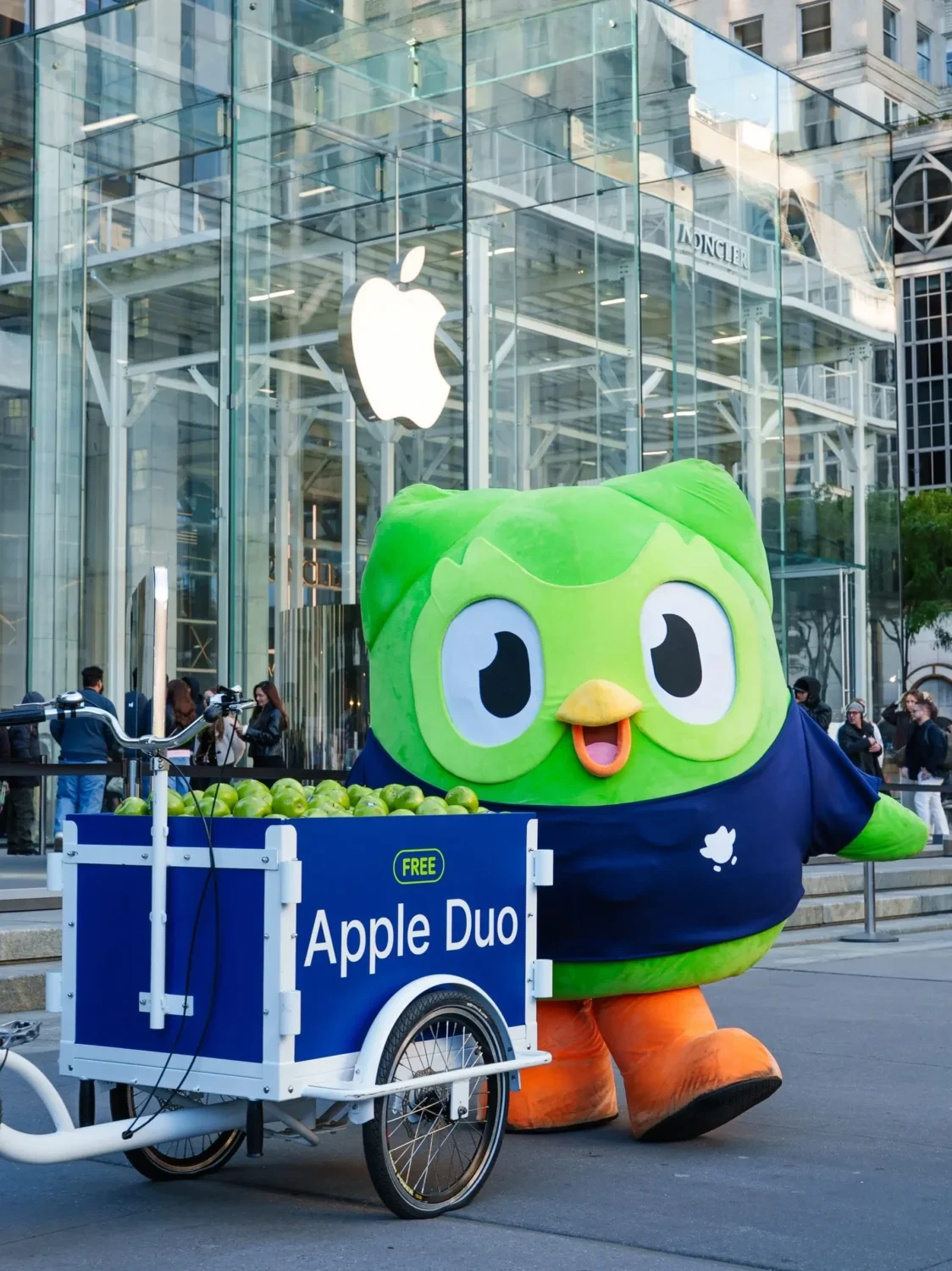 Mascota Duo de Duolingo junto a una bicicleta de carga Apple Duo frente a la tienda Apple Fifth Avenue en Nueva York.