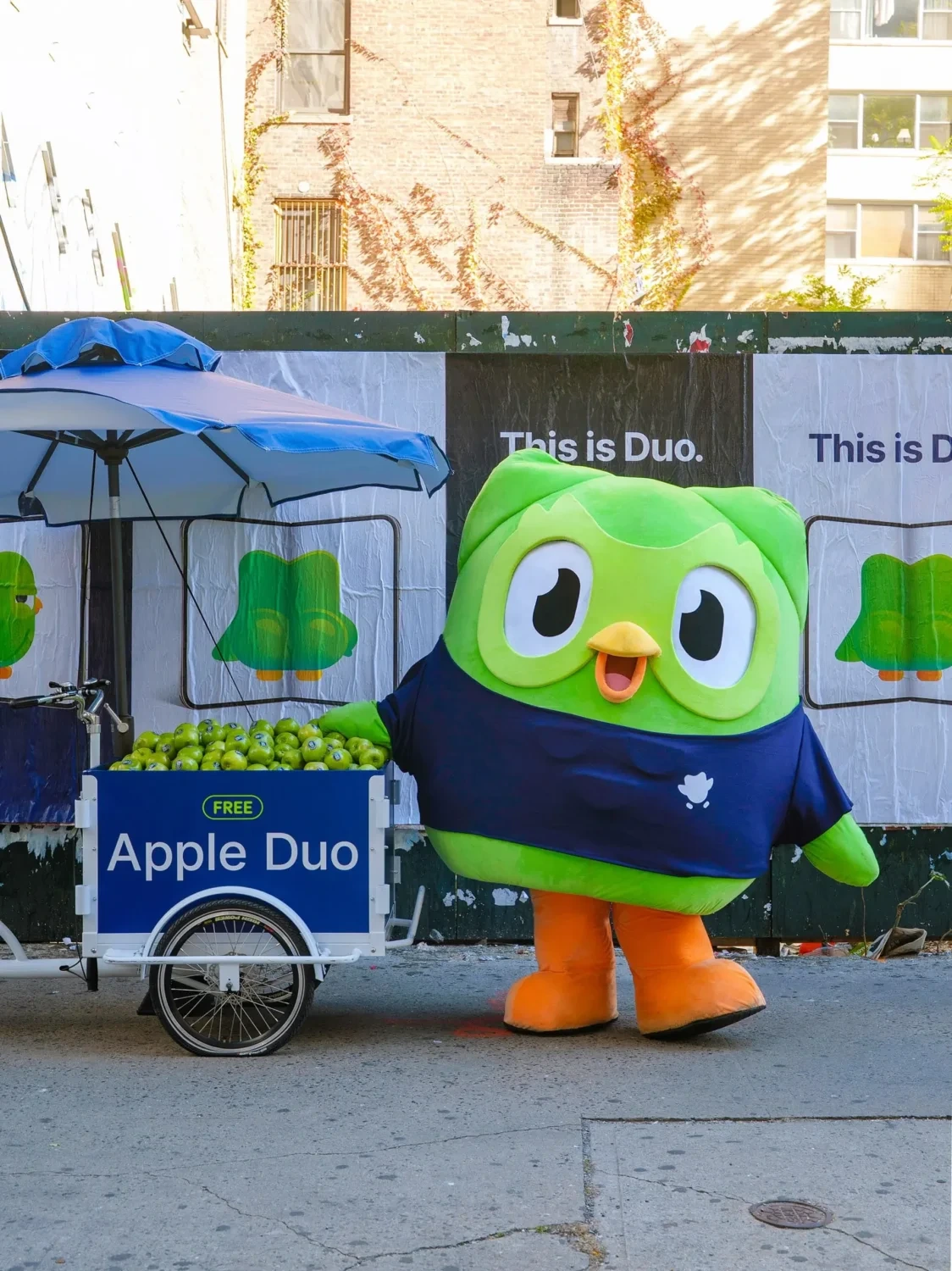 Duo de Duolingo junto a la bicicleta Apple Duo frente a carteles que dicen This is Duo en Nueva York.