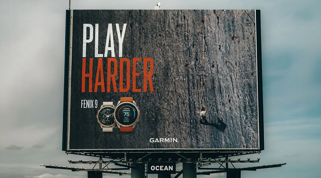 Billboard de Garmin Fenix 9 con el mensaje Play Harder y un escalador real ascendiendo la superficie del anuncio.