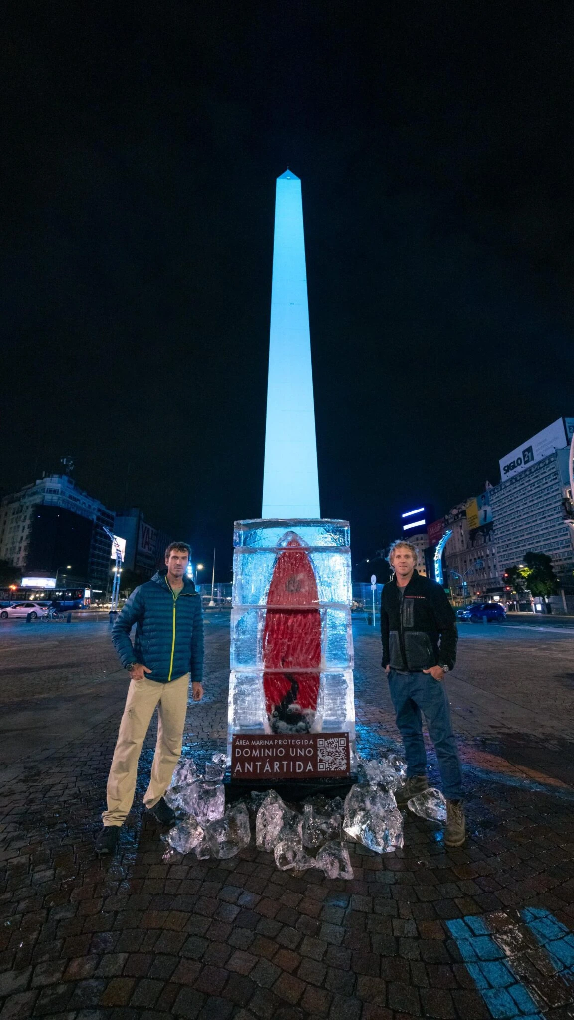 Joaquín y Julián Azulay junto a la tabla de surf congelada de Antártica - Dominio Uno con el Obelisco iluminado al fondo.