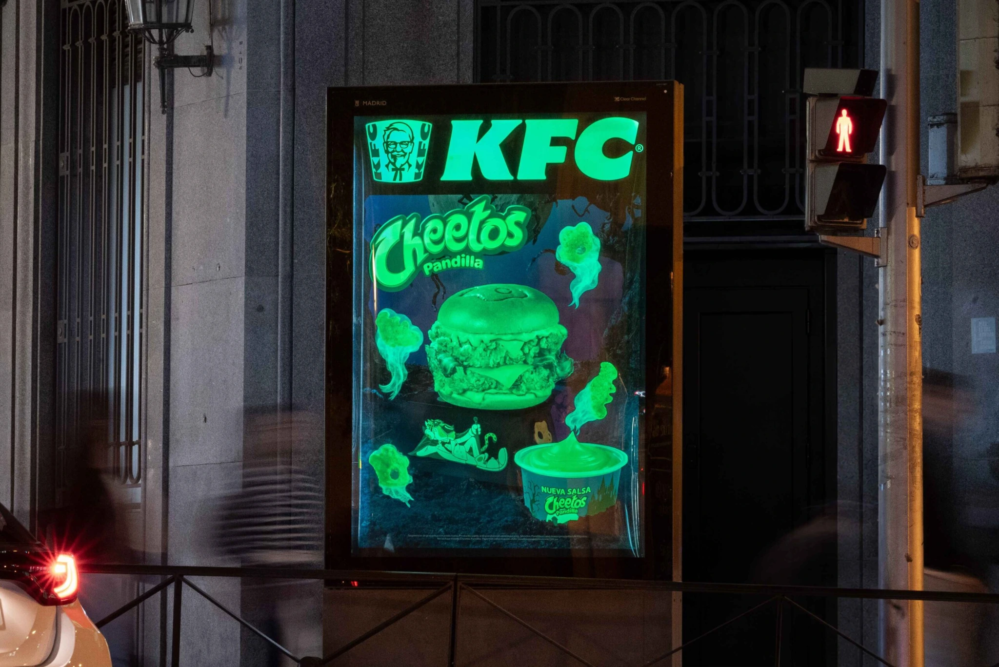 Mupi de KFC x Cheetos Pandilla iluminado por la noche en Madrid, con fantasmas y elementos luminiscentes de Halloween.