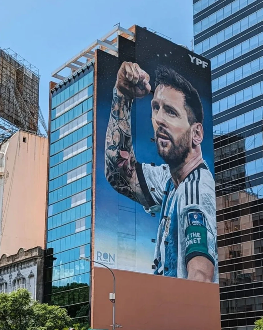 Mural monumental de Lionel Messi con camiseta de Argentina y el brazo levantado sobre la fachada de un edificio.
