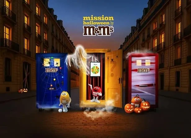 Visual promocional de M&M’s Mission Halloween con tres puertas temáticas y personajes de la marca en una calle iluminada al atardecer.