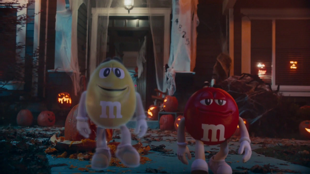 Personajes amarillo y rojo de M&M’s caminando en una calle decorada para Halloween con calabazas encendidas y ambiente nocturno.