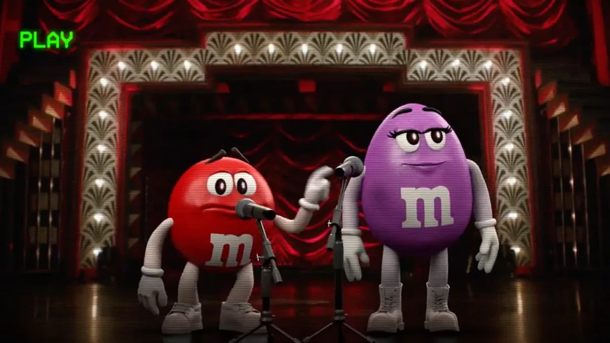 Personajes rojo y morado de M&M’s en un escenario con micrófonos y telones rojos dentro de una escena promocional de Halloween.