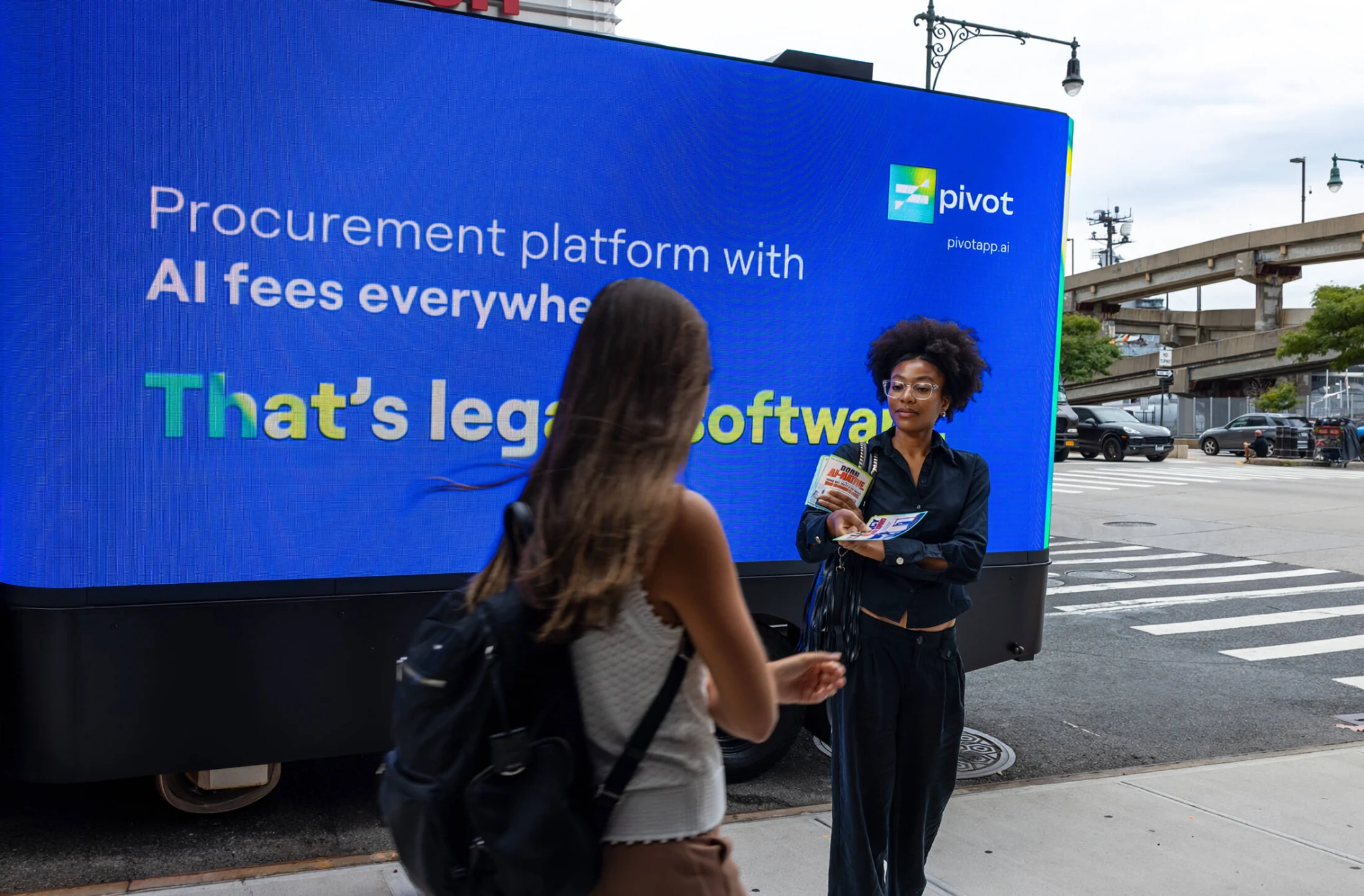 Camión con pantalla digital de Pivot en Nueva York mostrando un mensaje sobre tarifas de IA y software legacy mientras una activación de calle entrega material a peatones.