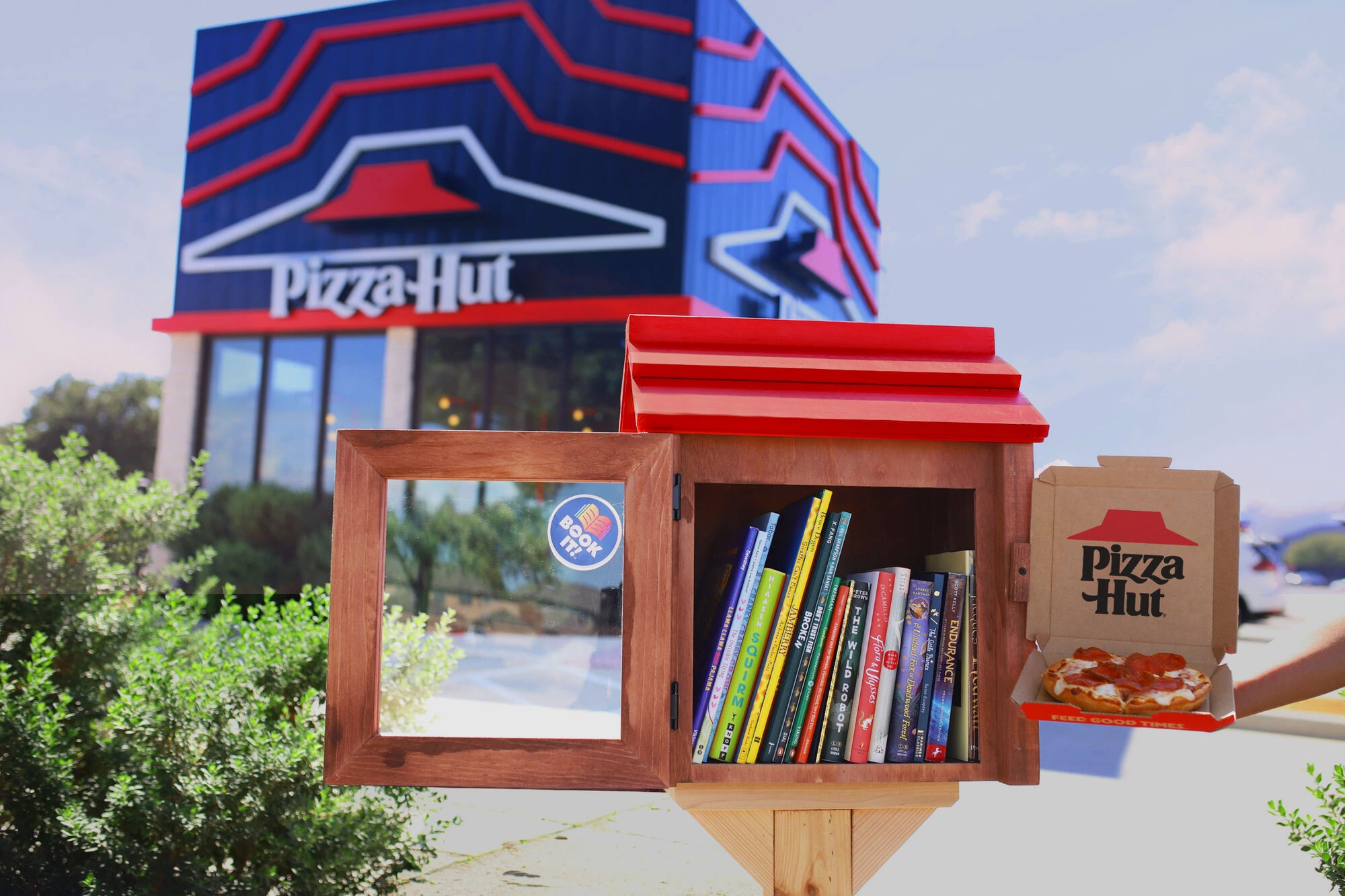 Little Hut Library abierta frente a un restaurante Pizza Hut, mostrando libros en su interior y una caja de pizza al costado.