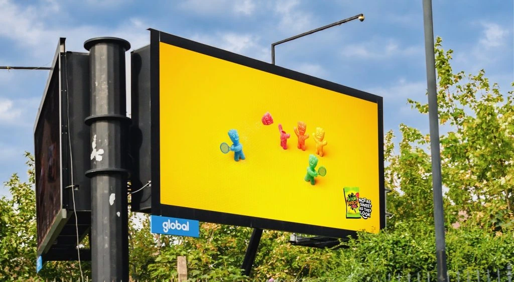 Billboard digital de Sour Patch Kids con personajes de colores en una escena inspirada en un juego de tenis y el mensaje Not As Sweet As They Look.