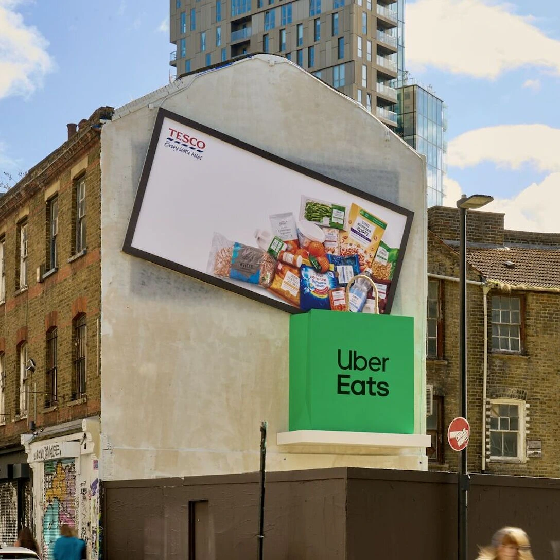 Billboard especial de Tesco inclinado sobre una gran bolsa verde de Uber Eats, simulando que la compra semanal cae dentro del delivery bag.