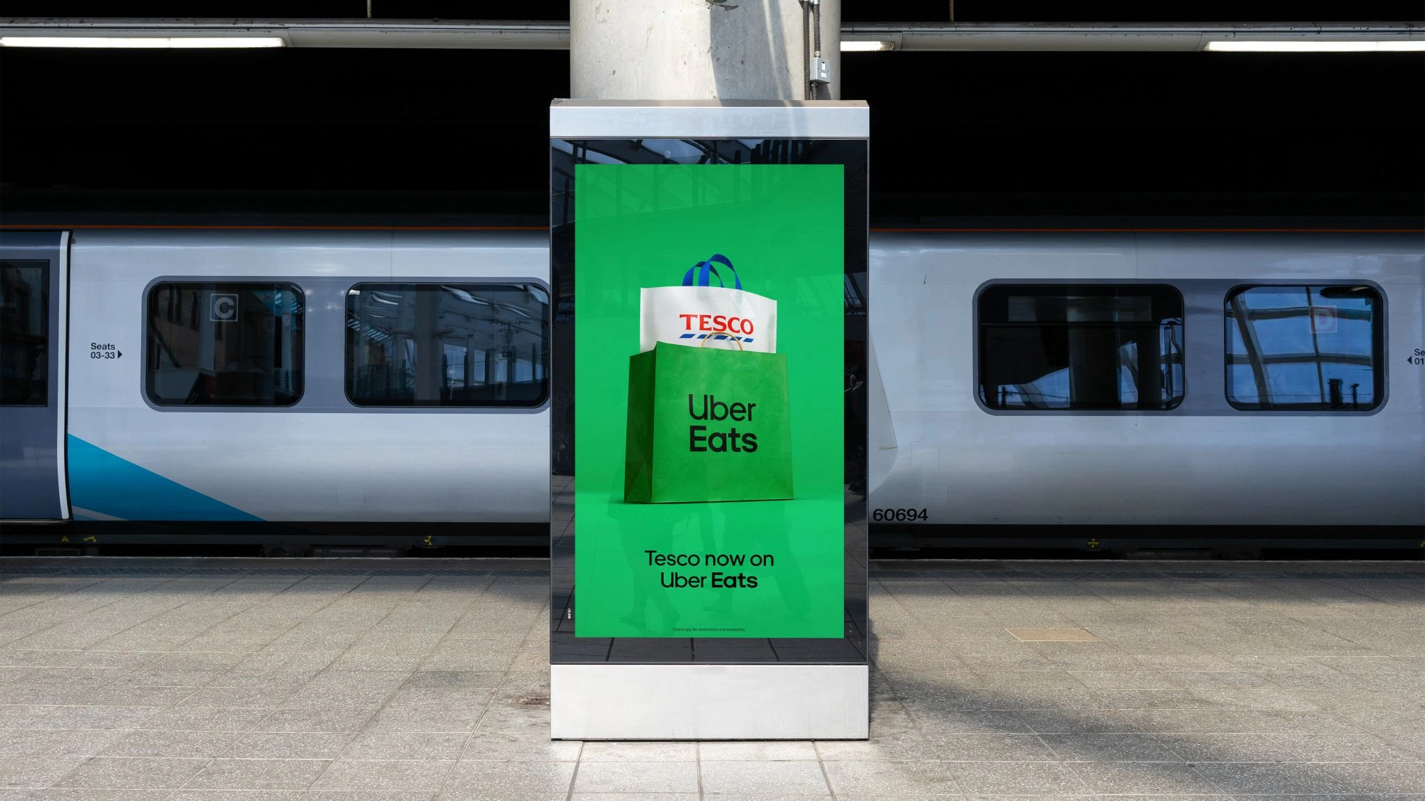 Pantalla digital en estación de tren con fondo verde mostrando una bolsa de Uber Eats con una bolsa de Tesco dentro y el mensaje Tesco now on Uber Eats.