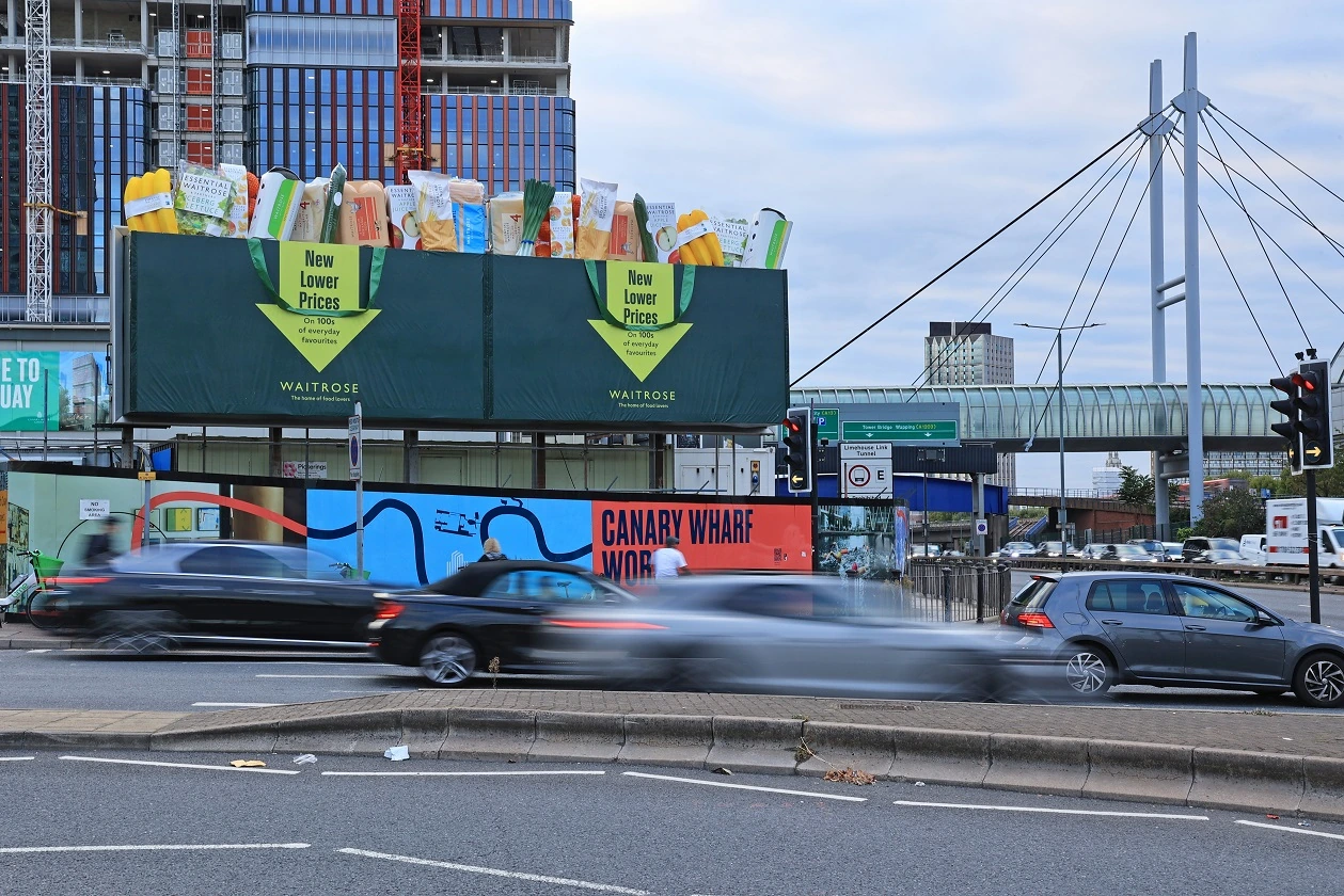 Instalación OOH de Waitrose en Aspen Way, Canary Wharf, con dos bolsas gigantes y productos sobresaliendo por encima de la estructura publicitaria.